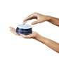 Hand holding Tidal Brightening Cream open jar with opposite hand touching light blue sugar like balm.